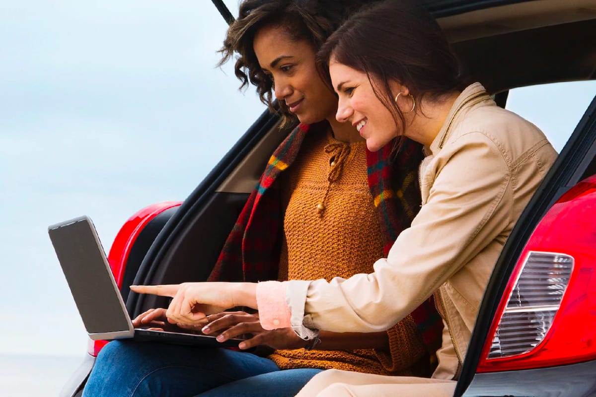 Two girls sitting on a car's trunk, while looking at a computer screen